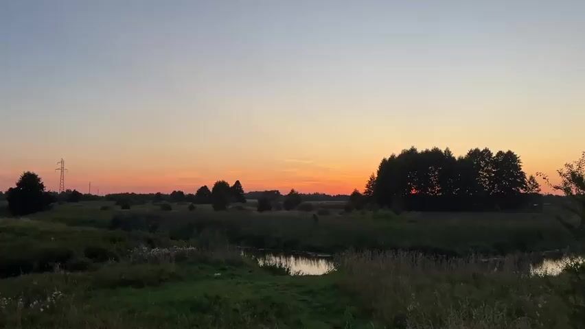 A sunset paints the sky with hues of orange and pink over a tranquil rural landscape. A small river winds through a field of tall grass and scattered trees, with a power line tower visible in the distance. The trees on the right form a dense silhouette against the vibrant sky. The overall impression is one of peaceful evening serenity in nature.