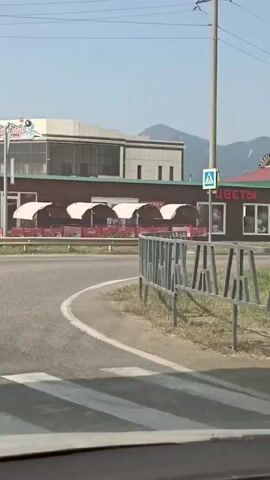 The video captures a view from a vehicle traveling along a paved road. On the right side of the road, a row of buildings is visible. The closest building is a small, dark wooden structure with a green roof and several windows. To its right, a slightly larger, darker wooden building with a red roof features a large metal barbecue grill outside. Further to the right, a more modern building with a white facade and a curved section is partially visible. A sign with mountains and a circular logo is mounted on top of this modern building. The buildings appear to be commercial establishments, with some signs indicating services like "REPAIR," "RENT," and "SALE." There are outdoor seating areas with red tables and white canopies in front of some of the establishments. The environment is semi-rural, with tall grass and some vegetation bordering the road and surrounding the buildings. Mountains are visible in the background under a clear blue sky. The camera appears to be mounted inside the vehicle, possibly on the dashboard or windshield, as the lower edge of the frame shows the vehicle's interior. The video is brief, showing the buildings as the vehicle passes them.