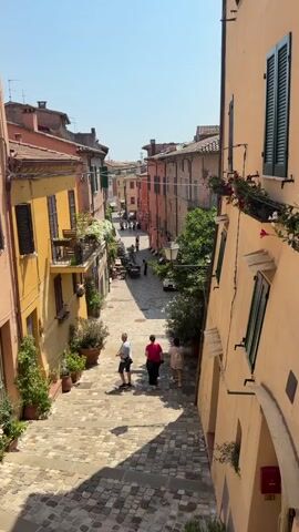 The video shows a narrow cobblestone street in an Italian town, lined with colorful buildings. The street slopes downwards, and there are steps leading down. Three people are walking down the street away from the camera. The person in front is wearing a white t-shirt and shorts. The person in the middle is wearing a red t-shirt and dark pants. The person in the back is wearing a light-colored dress. There are potted plants on the sides of the street and balconies with flowers on the buildings. The sky is clear and blue, indicating a sunny day.