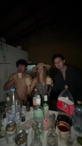 The video shows three young people, two males and one female, gathered around a table with drinks. The male on the left is shirtless and drinking from a mug. The female in the center is wearing a straw hat and a patterned top, and she is also drinking from a mug. The male on the right is wearing a dark collared shirt and is drinking from a mug. The table is cluttered with various bottles of alcohol and other drinks, as well as some plastic bags. The lighting is dim, suggesting it is nighttime or indoors with low light. The background appears to be a room with a slanted ceiling and some hanging plants on the left. The overall impression is a casual gathering or party.