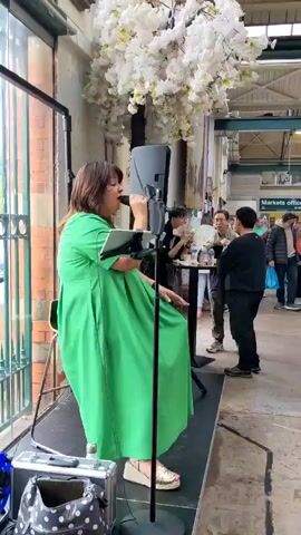 The video begins with a woman in a green dress singing into a microphone on a stage. The stage is set up in what appears to be a market or indoor food court, with stalls and shoppers visible in the background. The setting is decorated with white artificial flowers and string lights. The camera pans to the right, showing various stalls, including one for coffee and tea. Shoppers are seen browsing and sitting at tables. The video continues to pan, revealing more of the market, with signs for "Crepes" and "Food Court" visible. The overall atmosphere appears to be lively and bustling.