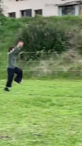 In a full shot, a young man in a green shirt and black pants performs a cartwheel in a grassy yard. The backdrop includes a stone house partially obscured by a grassy hill and a bare tree. A wire fence stretches across the middle ground. After executing the cartwheel, the man stumbles slightly, then regains his balance.
