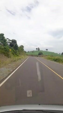 The video depicts a view from a moving car on a paved road with a yellow line on one side and a white line on the other. The sky is overcast. On the left, there is a grassy hillside with trees and a road sign. On the right, a grassy hill with a few trees is visible in the distance. A motorcyclist rides in the distance on the road ahead. The windshield of the car is visible at the bottom of the frame.  Sounds of people talking are audible.