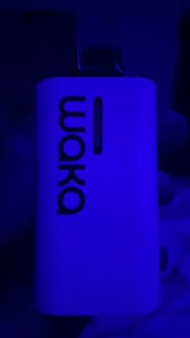 The video shows a close-up, handheld shot of a white rectangular electronic device with the black logo "waka" printed vertically on its side. The device is illuminated by a blue light, casting a blue hue over its surface and the surrounding dark environment. A person's hands are visible, holding the device. The device appears to be a disposable vape. The shot is slightly shaky, suggesting movement.