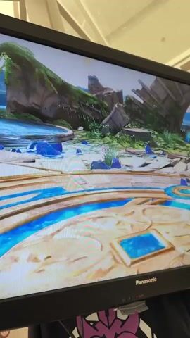 A person records a video of a computer monitor. The monitor displays a video game featuring Pikachu, a yellow mouse-like creature with black-tipped ears and a lightning-bolt shaped tail. The game environments change throughout the clip, showing different stages and backgrounds. The monitor itself is black with a Panasonic logo on the bottom right. The background behind the monitor is out of focus but appears to be an indoor setting with a light-colored ceiling and some cables or wires visible.