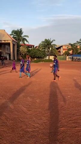 Several people are walking on a red dirt field. Most of them are wearing soccer uniforms. The uniforms are blue with red and white accents. Some palm trees are in the background, as well as several buildings. One of the people is carrying a soccer ball.