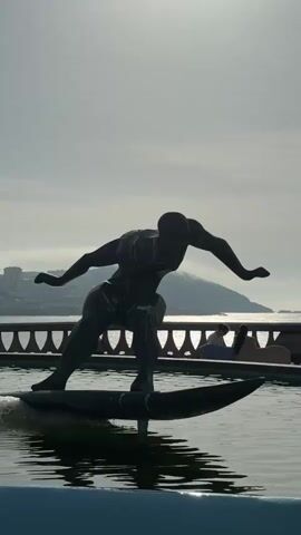 A statue of a surfer on a surfboard is in the foreground, with water splashing around its base. In the background, there is a railing, a person sitting on a bench, a body of water, and a hill or mountain in the distance. The sky is overcast. A bird flies across the sky.