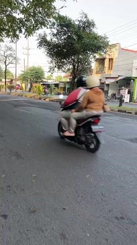 The video shows a street scene with several motorcycles. In the foreground, on the left, a motorcycle is moving away from the camera, with two people on it. The rider is wearing a brown jacket, and the passenger is wearing a white helmet. The motorcycle is dark in color. The road is asphalt and appears wet in places. In the background, several other motorcycles are parked or moving. There are buildings on the right side of the street, with awnings extending over storefronts. One building has a red roof. Trees and utility poles are visible on the left side of the street. The lighting suggests it is daytime, possibly late afternoon or early morning, with soft shadows.