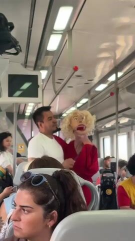 A man in a white shirt is dancing with a blonde wig wearing a red dress on a public transport vehicle. Other passengers are visible in the background and foreground. The interior of the vehicle appears to be a train or bus, with overhead lights and handrails. The focus is on the man and the puppet-like figure he is dancing with.