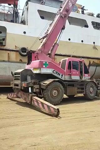 A pink and grey mobile crane, labeled "CRN 1" and "PANTHER", is positioned next to the hull of a large, yellow and white ship. The crane's boom is raised, and it appears to be lifting or lowering two white spherical buoys. A small red truck with a flatbed is parked behind the crane, and two men are visible in the back of the truck. Another man, wearing a hard hat, stands near the rear of the truck, looking towards the buoys. The ground is covered in dirt and sand. In the background, another ship, primarily blue and white, is partially visible. The sky is bright and overcast, suggesting daytime.