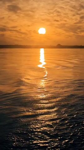 A vertical video displays a sunset over a body of water. The sun, a bright orange orb, is low in the sky, partially obscured by wispy clouds. Its reflection creates a shimmering, golden path across the water's surface. The water itself is dark in the foreground, with visible ripples and waves, transitioning to a lighter, more reflective orange hue closer to the horizon. A distant, dark landmass or silhouetted structure is visible on the horizon line. The overall impression is one of a serene and warm natural landscape.