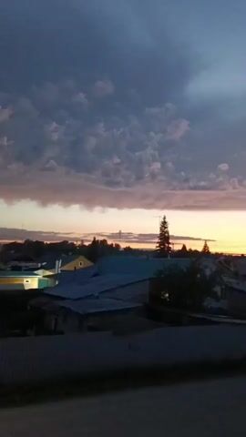 The video shows a rural landscape at dusk. The sky is filled with dramatic clouds in shades of purple, gray, and orange, indicating the setting sun. Below the sky, there are several houses with dark roofs, and a tall, slender tree stands out against the horizon. Some of the houses have lights on. In the foreground, a dark, possibly paved surface and a fence are visible, suggesting the viewpoint is from a street or road looking towards the houses. The overall atmosphere is calm and serene, characteristic of the end of the day.