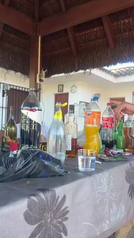 Here is a detailed description of the video:

The video begins with a close-up of a table covered with a floral-patterned tablecloth, littered with various bottles of alcoholic beverages, soft drinks, a glass with liquid, and other items like cigarettes and a lighter. A large black bag is also on the table.

The camera then pulls back to reveal a thatched roof structure, presumably a gazebo or covered patio. Underneath, three men are seated around the table. One man is partially visible behind the table, another is seated on the left side of the table, wearing sunglasses, and a third is seated to the right.

The camera shifts, blurring as it moves, showing the seated men from different angles. Beyond the structure, a glimpse of a white building with a red door and a manicured green lawn are seen, suggesting an outdoor setting, likely a backyard or patio. The general impression is of a casual gathering, possibly a party or a relaxed hangout, with alcoholic beverages and drinks.