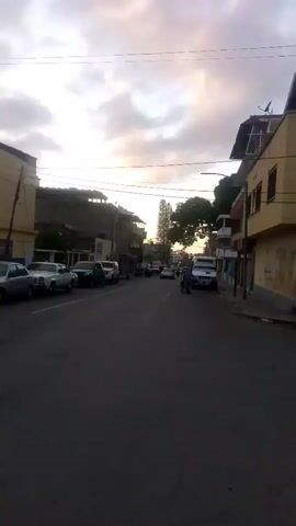 The video shows a street scene at dusk. Cars, motorcycles, and a white van are on the road. A person is pushing a cart. Buildings line the street, and trees are visible. The sky is cloudy with the sun setting.