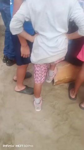 The video shows a group of people on a sandy surface, possibly a beach. In the foreground, a person wearing a white long-sleeved shirt and pink patterned shorts is partially visible, facing away from the camera. To their right, another person in pink shorts is also visible from the waist down. The central focus of the video is a gathering of individuals seated or kneeling. Two people are actively playing drums. One drum, a conga-like instrument, is brown and cylindrical. Another drum appears to be a bongo or djembe, white on top and possibly made of wood. One person, wearing a white t-shirt with an image of a woman on it, is playing the white-topped drum. Another person, wearing a red cap, is also near the drums. A woman in a white top is in the background, holding a conch shell to her mouth as if to blow it, and appears to be singing or shouting. Several other individuals are present, observing the musicians and the woman with the shell. The environment appears to be outdoors, with sand visible underfoot and a weathered wooden post or structure in the background. The overall atmosphere suggests a lively, possibly celebratory or musical gathering.