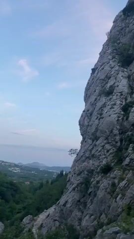 The video captures a dramatic natural landscape in Zupci, Montenegro. A very steep and rugged grey and white rocky mountainside, partially covered with lush green trees and bushes, dominates the frame. The camera slowly pans downwards and to the right, revealing more of the intricate rock formations and vegetation. In the mid-ground, a small, dark figure, appearing to be a person, can be seen on the rocky slope, possibly hiking or climbing.

Above the mountain, the sky is a soft blue with scattered wispy clouds. In the distant background, a verdant valley stretches out, leading towards what appears to be a large body of water, likely the sea, under a lighter horizon. The lighting suggests it is daytime, possibly late afternoon or early morning, given the gentle illumination. There is no visible text in the video. Overlapping voices in the background, speaking in what sounds like Russian, express excitement or urgency; one voice states, "This needs to be posted, guys. Urgently. This is me, oh my. That's it, everyone," suggesting an engaging and memorable experience in this natural setting.