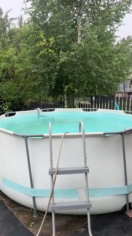 The video shows a backyard swimming pool on a cloudy day. Raindrops are visible on the surface of the pool and the surrounding ground. A ladder is positioned next to the pool, with a hose draped over it. In the background, there are trees, a wooden fence, and a white car parked on the grass. A wooden walkway is visible on the left side of the frame.