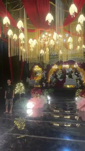 The video shows an elaborately decorated event space, likely for a wedding or formal celebration. The scene is set at night, with abundant artificial lighting. A person is standing on the left side of the frame, wearing a dark t-shirt and shorts, looking towards the stage area. The stage itself is a focal point, adorned with a large floral arrangement, a cake, and seating furniture. Above the stage, a grand chandelier hangs amidst draped red fabric and numerous hanging lights. A pathway, possibly a red carpet or a reflective surface, leads towards the stage, flanked by decorative trees with white lights and large spherical lights. The overall atmosphere is festive and luxurious, with a clear emphasis on visual spectacle through lighting and decor.