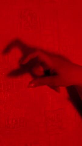 A red-lit video features a hand casting a shadow on a textured red background. The fingers of the hand are arched to create a "monster claw" shadow. The hand is female, with red nails. A voice in the background says something akin to a spooky phrase.