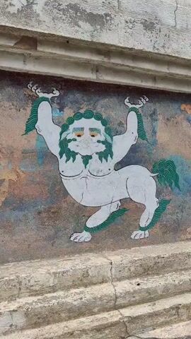 The video shows a close-up view of a painted mural on the side of a stupa. The mural depicts a mythical creature that appears to be a lion with human-like features, a beard, and green hair and tail. The creature is white with green accents and has human-like arms and chest. To the left of the creature, there is a circular symbol with intricate patterns and a blue and green center. The background of the mural is weathered and shows hints of other faded paintings. The stupa itself is made of stone and concrete, with visible cracks and signs of age. The top portion of the stupa is visible in the last frame, showing a conical structure with colorful bands. The sky is overcast.