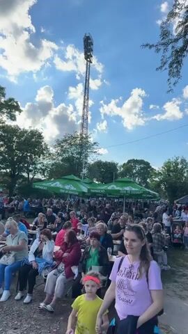 The video shows a crowded outdoor event with many people gathered. The scene is set under a blue sky with white clouds. Tall trees border the area, and a tall, slender tower with lights is visible in the background. In the foreground, people are seated at picnic tables under green umbrellas. Further back, a multitude of people are standing and walking around tents, suggesting a market or festival. Smoke is rising from an area behind the tents, possibly indicating food vendors or a bonfire. The overall impression is of a lively community gathering.
