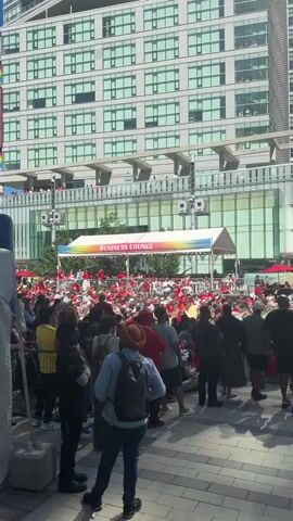 The video shows a large crowd of people gathered outdoors in front of a modern building with a glass facade. Many in the crowd are wearing red clothing. Above the crowd, a tent is set up with the words "BUSINESS LOUNGE" displayed on a colorful banner with a rainbow design. Small flags are also strung along the top of the tent. People are standing and sitting, and some appear to be looking towards the tent area where several individuals are visible, some standing and interacting. The overall atmosphere suggests an event or gathering.