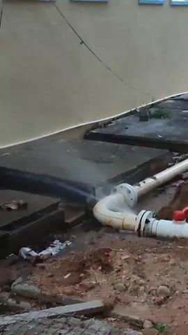 The video shows a damaged pipe expelling steam into the air. The pipes are white and cream-colored, with bolted connections. A red-handled valve is visible on one of the pipes. The pipe is located in a dirt area, partially buried, next to concrete slabs and a light beige wall with small windows. There is debris scattered around the pipe. The steam is obscuring the immediate area of the break.