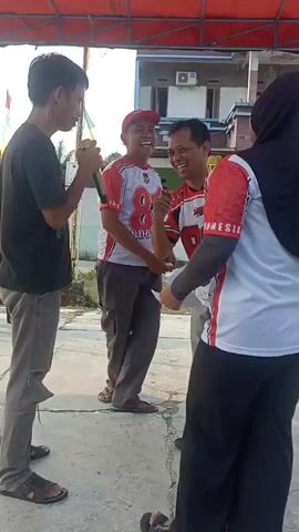 Four people are standing in an outdoor area under a red awning. A man in a black t-shirt and ripped grey pants holds a microphone. To his right, a man in a red and white shirt with the number 87 on it and a red baseball cap smiles and laughs. To his right, a person wearing a black hijab and a red and white shirt stands facing away from the camera, with their arm extended towards the man with the number 87 on his shirt. Another person, mostly out of frame, is partially visible between the man in the red and white shirt and the person in the hijab. The ground is paved. In the background, there is a building with windows and an air conditioning unit, and some flags on poles.