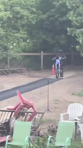 The video shows a person on a dirt bike performing a motocross stunt. The person is wearing a helmet and protective gear. The dirt bike is blue and white. The stunt involves jumping off a ramp and performing a flip in the air. The landing area is an inflatable airbag that reads "AIRBAGS". The environment is an outdoor setting with trees and a clear blue sky. There are also some chairs and debris in the foreground.