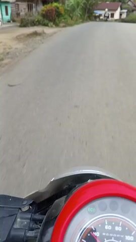 The video shows a point-of-view shot from someone driving a scooter down a narrow asphalt road in a rural area. The scooter's front panel, with a red accent, is visible at the bottom of the frame. Along the road are scattered houses of modest construction, surrounded by lush greenery including trees, shrubs, and other plants. In the background, a mountain range is partially obscured by an overcast sky. A red truck passes by in the distance along with another scooter. The scene is generally calm and appears to be taken during the daytime.