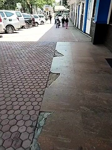 A person is walking on a tiled sidewalk. The tiles are broken in some places. There are cars parked on the side of the road. There are also buildings in the background.