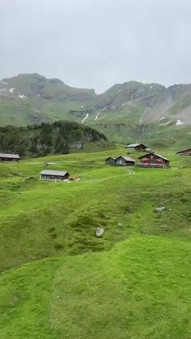 A wide shot captures a verdant mountainous landscape under an overcast sky. Several small, wooden buildings with red roofs are scattered across the rolling green hills. The terrain is lush and appears well-maintained, with patches of snow visible on the higher mountain slopes in the distance. The overall impression is one of a serene, rural mountain village or settlement.