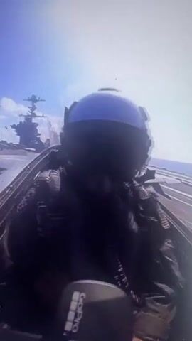 A first-person perspective video shows a pilot wearing a flight helmet and oxygen mask in the cockpit of a fighter jet. In the background, a large aircraft carrier is visible at sea under a bright sky. The camera is mounted on the pilot's helmet or in the cockpit, showing the pilot's hands on the controls and the instrument panel. The video appears to be taken during a pre-flight or taxiing phase on the carrier's deck.