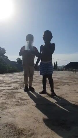 Two young boys stand on a concrete surface under a bright sun and clear blue sky. The boy on the left, wearing a t-shirt, pants, and dark shoes, holds a clear plastic container in his hands. The boy on the right, shirtless and wearing striped shorts, stands with his arms at his sides. Long shadows are cast on the ground by both boys. In the background, trees, buildings, and what appears to be a utility pole are visible. The overall environment suggests an outdoor setting on a sunny day.