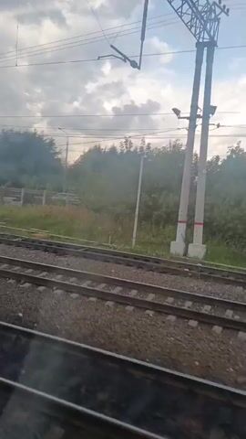 The video is filmed from inside a moving train, looking out a window. The train tracks are visible in the foreground. Beyond the tracks, there is a grassy area with trees and bushes. In the distance, there is a white truck parked near a fence. There are also power lines and utility poles visible in the scene. The sky is cloudy with patches of sunlight breaking through. The video appears to be taken during the daytime.