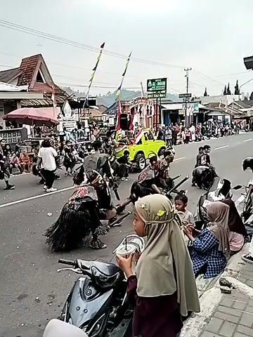 The video shows a street parade or cultural procession. People are dressed in elaborate costumes, some with headdresses, armor, and fringed skirts, and are holding props like swords and fans. Some individuals appear to be performing or dancing. Spectators are lined up along the side of the road, some sitting on motorcycles, watching the event. There are buildings, shops, and tents lining the street, suggesting an urban or town setting. Flags are visible, including what appear to be Indonesian flags. A bright yellow vehicle, possibly a jeep, is also part of the procession. The sky is overcast, and the overall atmosphere seems festive and communal.