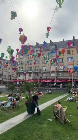 The video shows a public square decorated with numerous colorful hot air balloon-shaped ornaments hanging from wires strung between buildings. The sun is visible in the sky, partially obscured by clouds. In the foreground, a lush green lawn is interspersed with pathways. Several people are relaxing on lounge chairs spread across the lawn. In the middle ground, a man in a black t-shirt and blue jeans is leaning against a large potted plant. Behind the lawn and people, there is a row of buildings with windows and balconies, many of which have red awnings over ground-level establishments, likely cafes or restaurants. People can be seen sitting at tables under these awnings. The overall atmosphere appears to be one of a leisurely outdoor gathering or event.