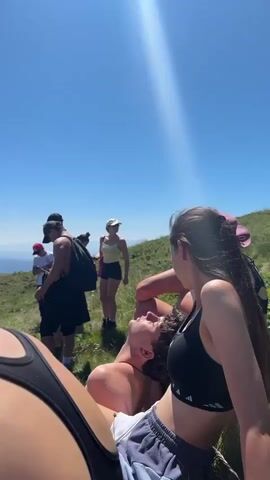 A group of people are gathered on a grassy hillside under a clear blue sky. Some are standing, some are sitting, and one person appears to be lying down. The people are dressed casually, in t-shirts, shorts, and tank tops, suggesting warm weather. Some are wearing hats and backpacks. The landscape appears to be mountainous, with trees visible in the distance. The overall atmosphere seems relaxed, possibly during a hike or a break on an outdoor excursion.