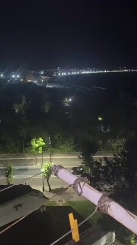The video shows a nighttime view from a balcony overlooking a city and the sea. The foreground features a clothesline with a yellow clothespin, and a white pipe secured with wire. Beyond the balcony, there are trees and a road with streetlights. In the distance, the city lights twinkle along the coastline, and the dark expanse of the sea is visible. The sky is dark and clear.