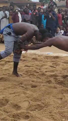 The video shows two shirtless men wrestling on a sand-covered ground. A crowd of people surrounds them, some recording the match with their phones. One wrestler wears blue shorts with a red drawstring, while the other is wearing gray pants with a blue cloth tied around his waist. The wrestling is intense, with the two men locked in a close struggle for dominance. The background is filled with onlookers, a building, and some trees, creating a lively outdoor setting for the wrestling match.