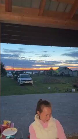 The video shows a sunset over a rural landscape. In the foreground, a person is partially visible. In the middle ground, a white car is parked on a grassy area. Behind the car, there is a fence and some buildings. The sky is filled with colorful clouds, and the sun is low on the horizon, casting a warm glow over the scene.