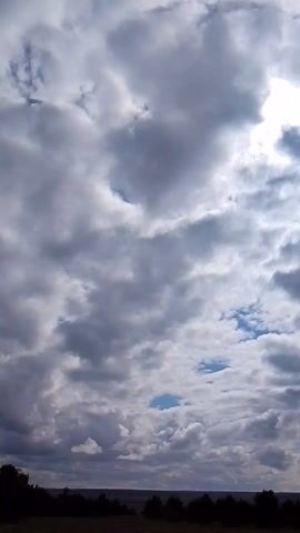 The video captures a dynamic, vertically oriented view of a cloudy daytime sky. Thick, predominantly grey and white clouds continuously shift and move across the frame. Bright sunlight periodically breaks through the cloud cover, creating luminous patches that suggest the sun's presence behind the clouds. Intermittent glimpses of clear blue sky are also visible between the cloud formations. Dark, indistinct shapes, likely tree lines, are visible along the far right edge of the frame, hinting at a distant horizon. The activity shown is the natural movement of clouds across the sky, possibly accelerated by a time-lapse effect given their rapid change. The weather appears to be partly cloudy or overcast with breaks of sun. No people, distinct objects, or visible text are present in the video. The provided location context is Елец-Маланино, Russia.