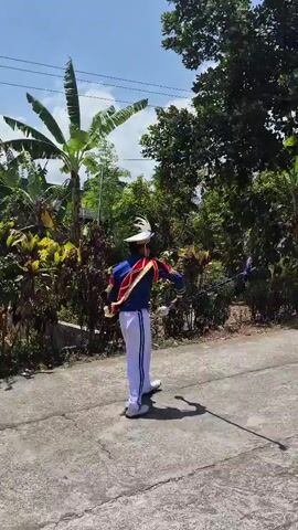 A person in a blue and red marching band uniform with white pants and boots and a white hat with a plume is performing with a baton in an outdoor street setting. The person is twirling the baton, which has a red tip. Behind the person, there is a house with a tiled roof and a green wall. There are also trees and plants in the background. The sky is blue with some clouds. Another person in a similar uniform is visible to the right. The video is shaky and appears to be filmed on a mobile device.