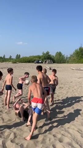 The video shows a group of young boys playing on a sandy beach. The sky is clear and blue, and there are some trees and bushes in the background. The boys are all wearing swim trunks and appear to be engaged in a game that involves running and tackling. One boy is holding a ball, possibly a rugby ball, and is being chased by other boys. Another boy is on the ground, seemingly having been tackled. There are also some tents and vehicles visible in the distance, suggesting a campsite or picnic area. The overall scene depicts a lively and energetic outdoor activity.