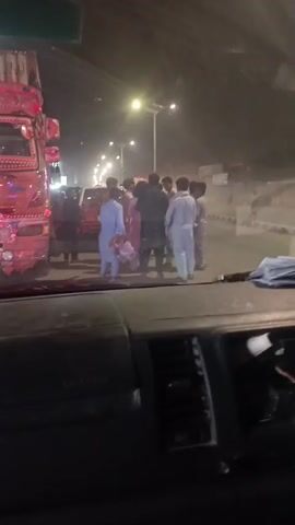 The video shows a group of people standing on a street at night. A large truck is parked on the left side of the frame. Several men and at least one boy are visible, wearing traditional Pakistani clothing. Some appear to be talking to each other. The scene is filmed from inside a vehicle. Streetlights illuminate the scene.
