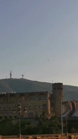 A static vertical shot captures a building in the foreground with a mountain and a cross in the background. The building appears to be made of concrete and has a Brutalist architectural style. It features a series of arched windows and a tall, cylindrical tower on its right side. The mountain behind the building is covered in vegetation, and at its summit, a large cross and a telecommunications tower are visible. The sky is clear and blue. A car is partially visible in the bottom left corner, suggesting a street or road in the immediate foreground. The overall lighting suggests it is daytime, possibly late afternoon or early morning due to the soft light. There are no people visible in the frame.