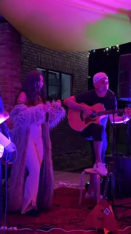 A man plays an acoustic guitar while standing on a stool. To his left, a woman in a long, furry coat and light-colored pants sings into a microphone. They are performing outdoors under a draped canopy illuminated by purple and green lights. In the foreground, red carpeting is visible, along with electrical wires and parts of musical equipment, including what appears to be a balalaika. The background shows a brick wall and a window. String lights are visible hanging from the canopy.