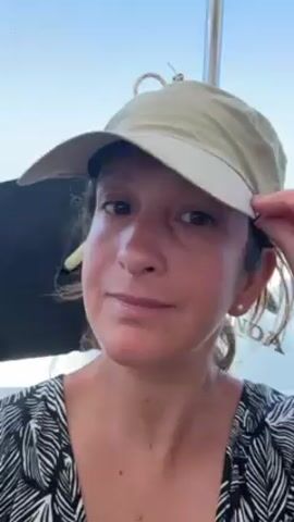The video shows a woman on a boat on the open water. She is wearing a black and white leaf-patterned top and a khaki baseball cap with a rope tied to the top. She touches the bill of her cap with her right hand and smiles at the camera. The background shows the water and wake from the boat, and an island is visible in the distance. The sky is blue.