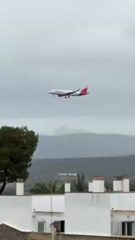 The video shows a passenger plane in the sky above a landscape. The plane is white with red and yellow markings and is descending, presumably for landing. The background includes trees, buildings, and distant mountains under a cloudy sky. The camera follows the plane's descent, adjusting its position to keep the aircraft in frame.
