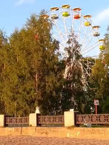 A Ferris wheel with colorful cabins is visible behind a row of trees and a decorative fence. The sky is blue with some clouds. The scene appears to be a park or an amusement area.